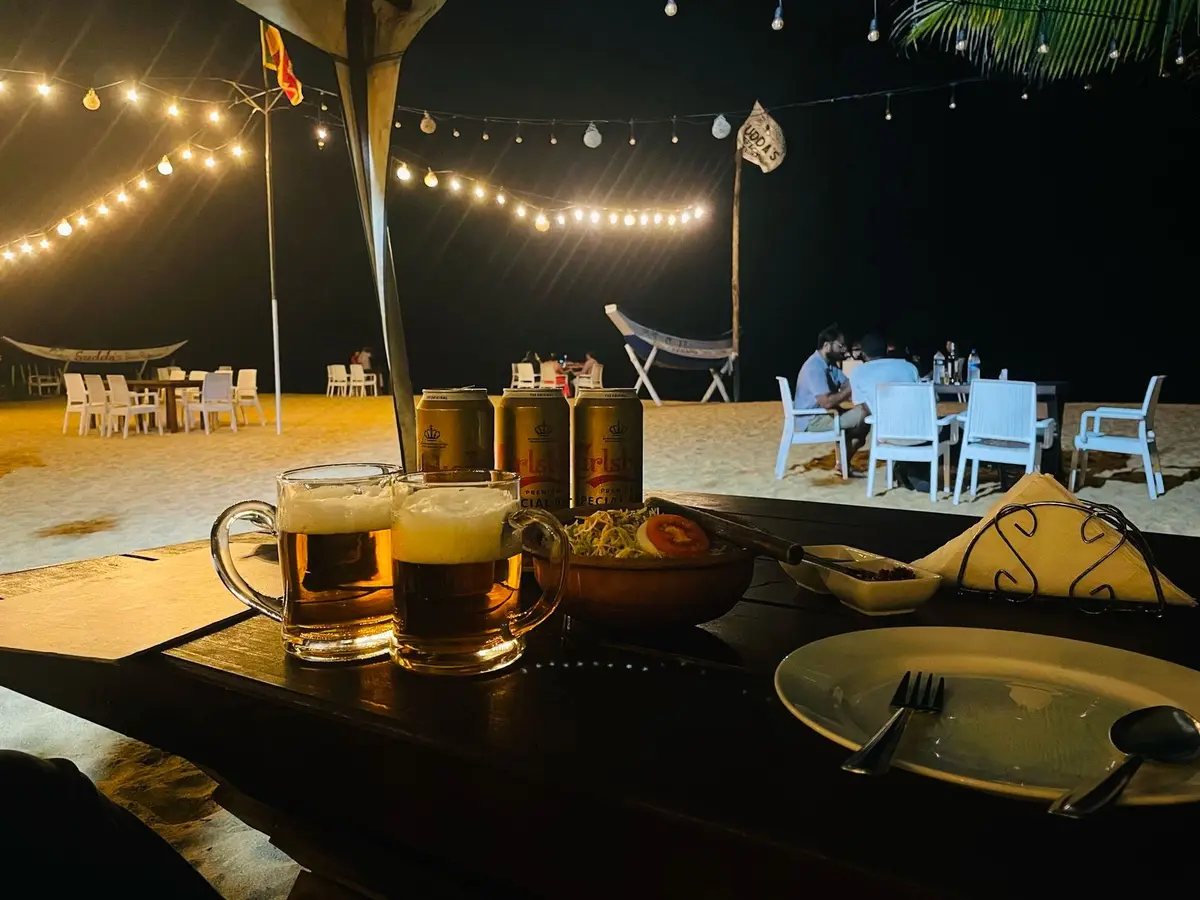 Tables on the sand under string lights at night with cold drinks and plates