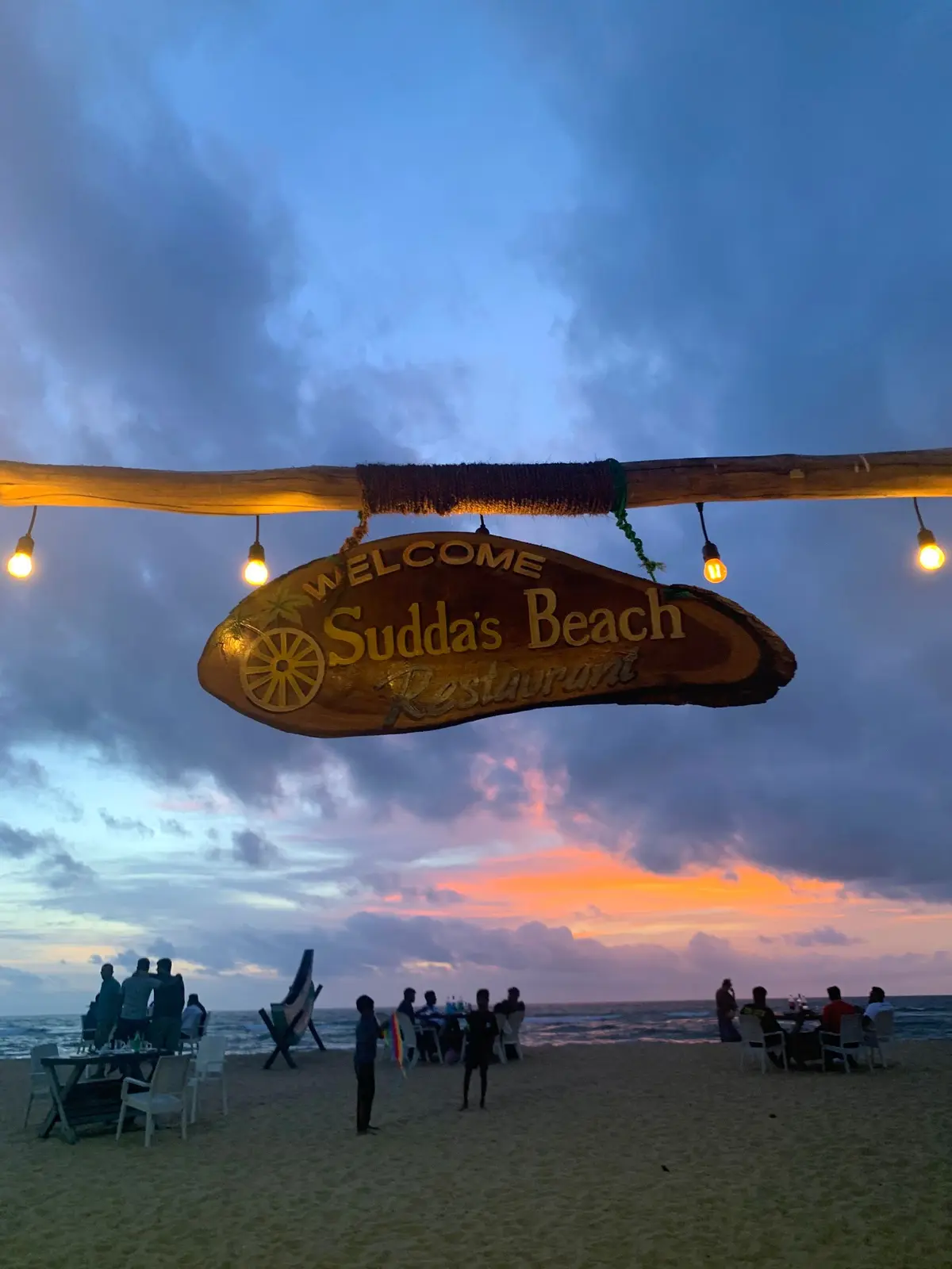 Carved wooden Sudda's Beach sign hanging under string lights at sunset, guests at tables on the beach