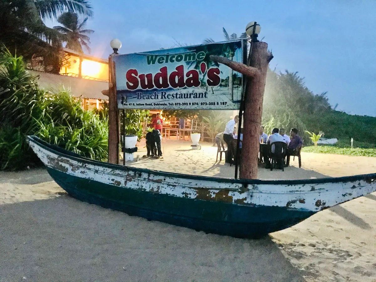 Welcome sign for Sudda's Beach Restaurant above an old fishing boat on the sand